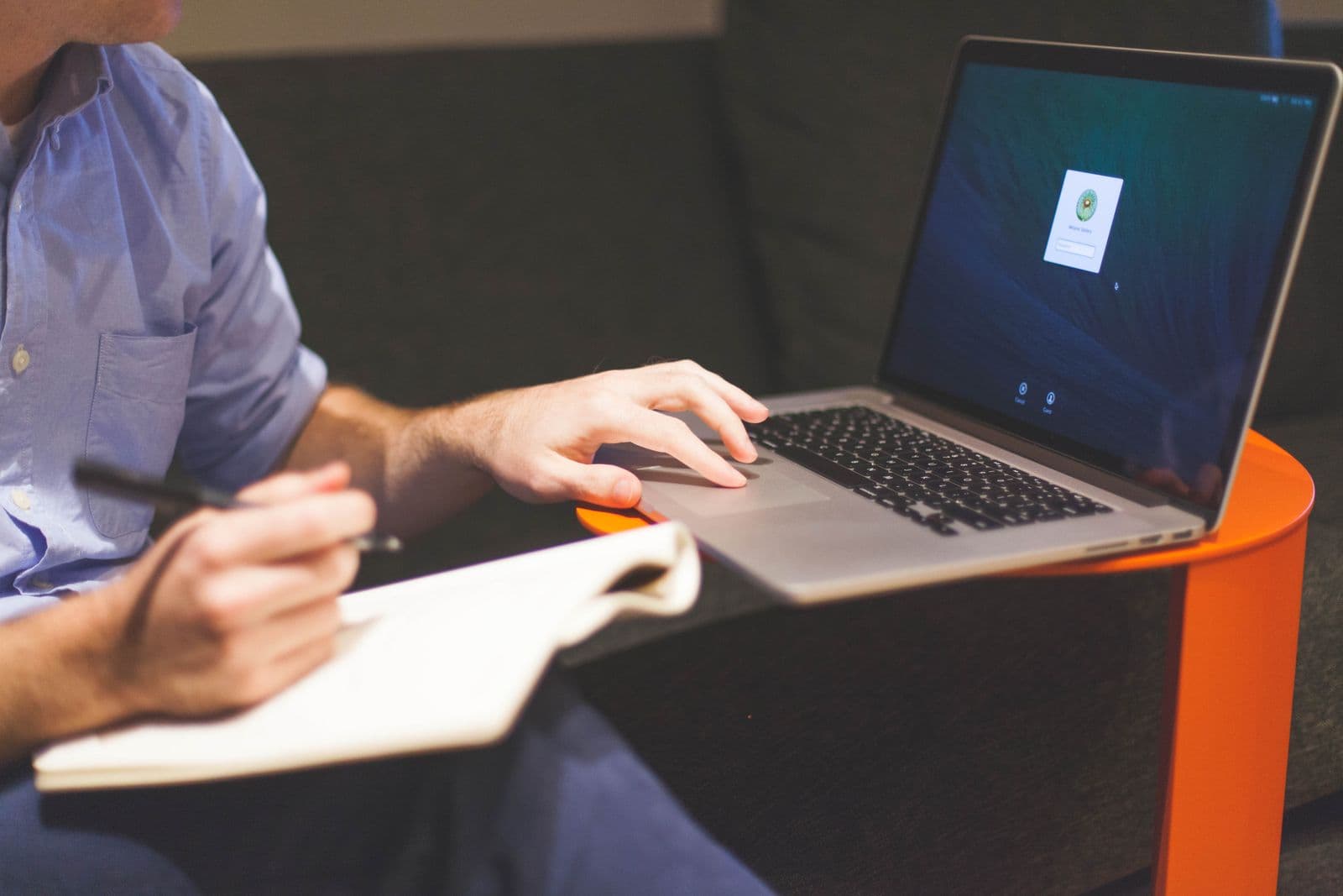 A person writing in an open notebook with one hand resting on the trackpad of a laptop