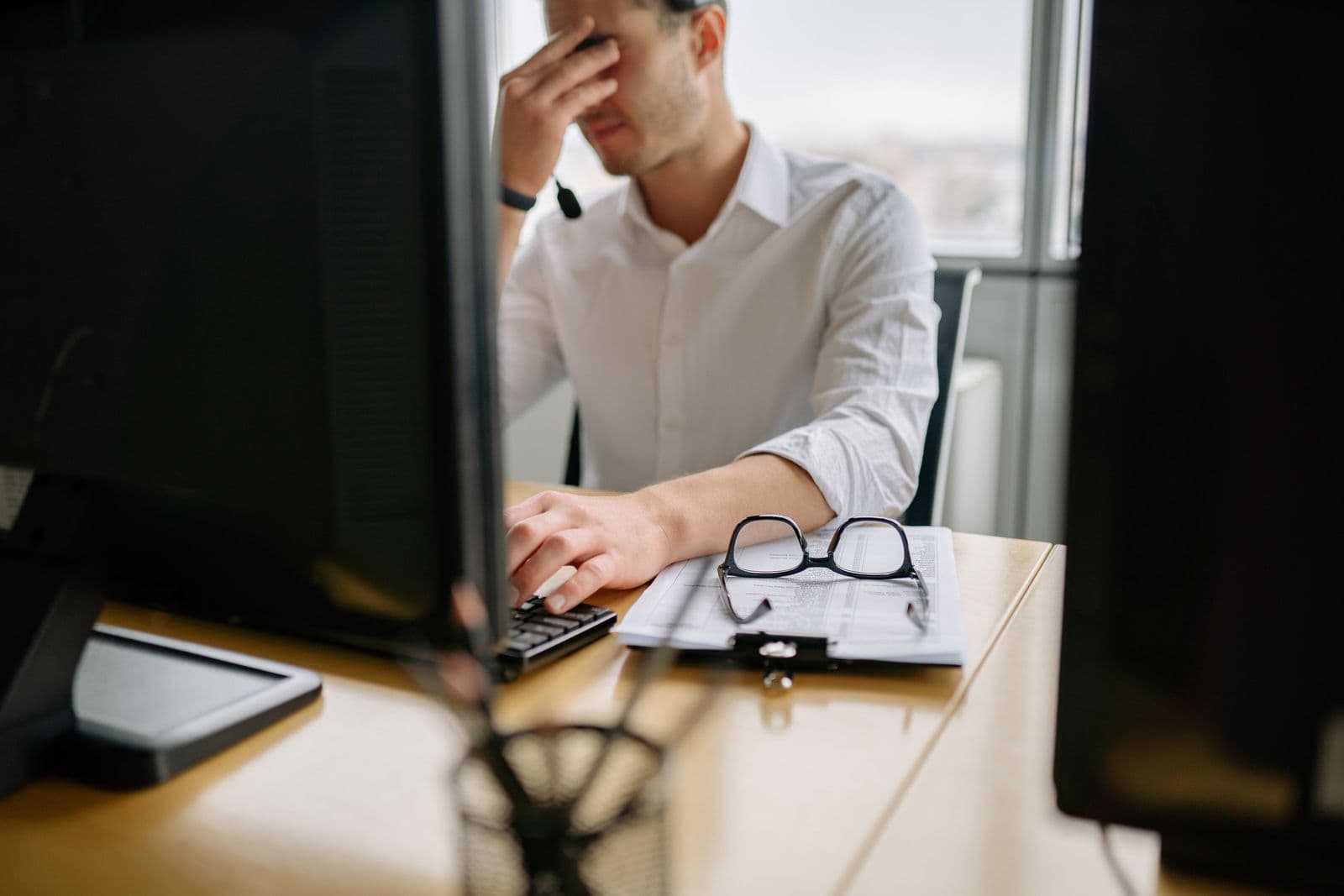 A man at a desk pinching the bridge of his nose, one hand on the keyboard, printed reports and glasses beside him